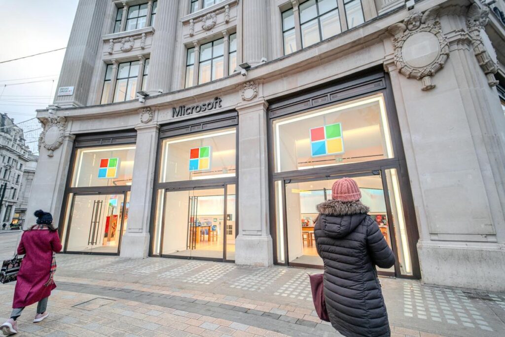 Pedestrians near illuminated Microsoft store facade at dusk.